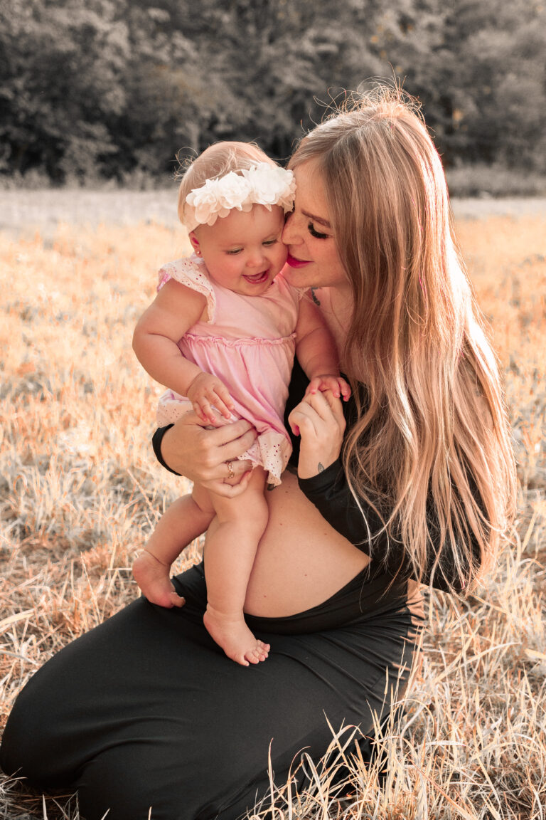Kleines Mädchen steht auf dem Schoß der schwangeren Mama auf Wiese in Fürth.