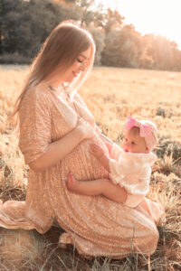 babybauch-fotos-fuerth Kleines Mädchen sitzt auf dem Schoß der schwangeren Mutter auf Wiese und hält ihren Babybauch fest in Fürth.