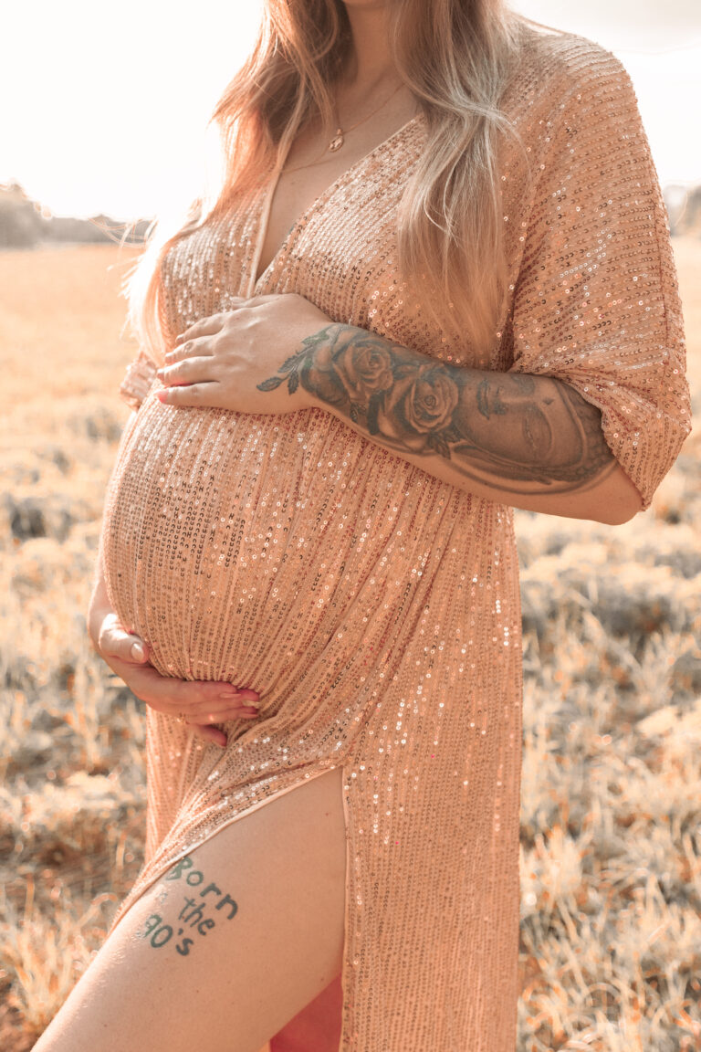 Schwangere Frau auf Wiese in Fürth trägt goldenes Kleid und hält ihren Babybauch mit beiden Händen fest.