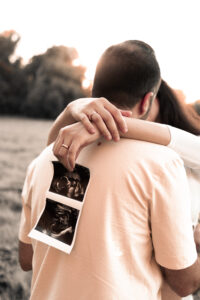 babybauch-shooting-paar-nuernberg ChatGPT: „Schwangere Frau hält Ultraschallbilder fest und umarmt ihren Mann auf Wiese in Nürnberg.