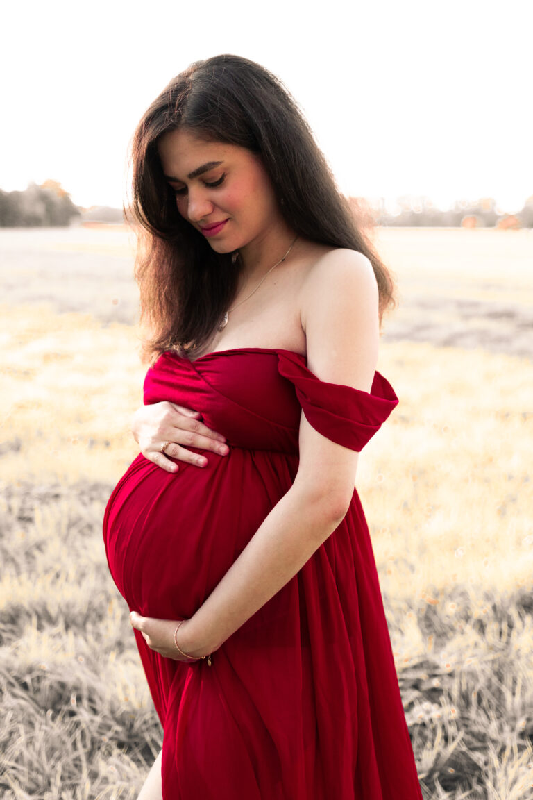Frau im roten Kleid hält Babybauch auf Wiese in Fürth und lächelt.