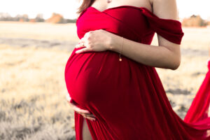emotionales-babybauchfoto-fuerth Schwangerschaftsfoto Frau im roten Kleid hält Babybauch auf Wiese in Fürth.
