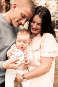 familienbilder-fuerth Frau hält ihr Baby im Arm, Mann spielt mit dem Baby auf Wiese.