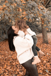 familienfotos-naturwiese-fuerth Frau hebt ihre Tochter vor Herbstbaum hoch und küsst sie auf Wiese.