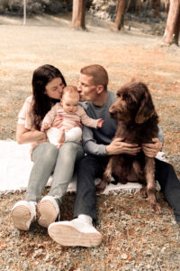 familienportraet-fuerth-natur Familie sitzt auf Decke im Park in Fürth, Frau hält und küsst Baby, Mann küsst Baby und hält Hund.