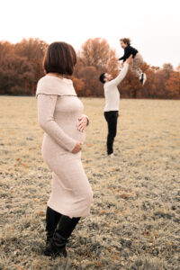 family-schwanger-shooting-nuernberg Schwangerschaftsfoto Familie auf Wiese in Nürnberg: Frau hält Babybauch, Mann spielt mit Sohn im Hintergrund.