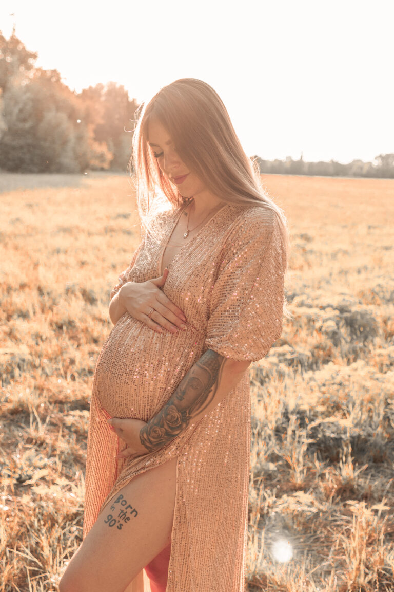 Blonde Frau auf Wiese in Fürth trägt goldenes Kleid und hält ihren Babybauch, lächelnd.
