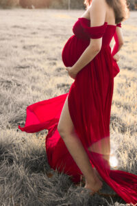 liebevolles-schwangerschaftsfoto-fuerth Schwangere Frau im roten Kleid steht auf einer Wiese in Fürth und hält ihren Babybauch fest.