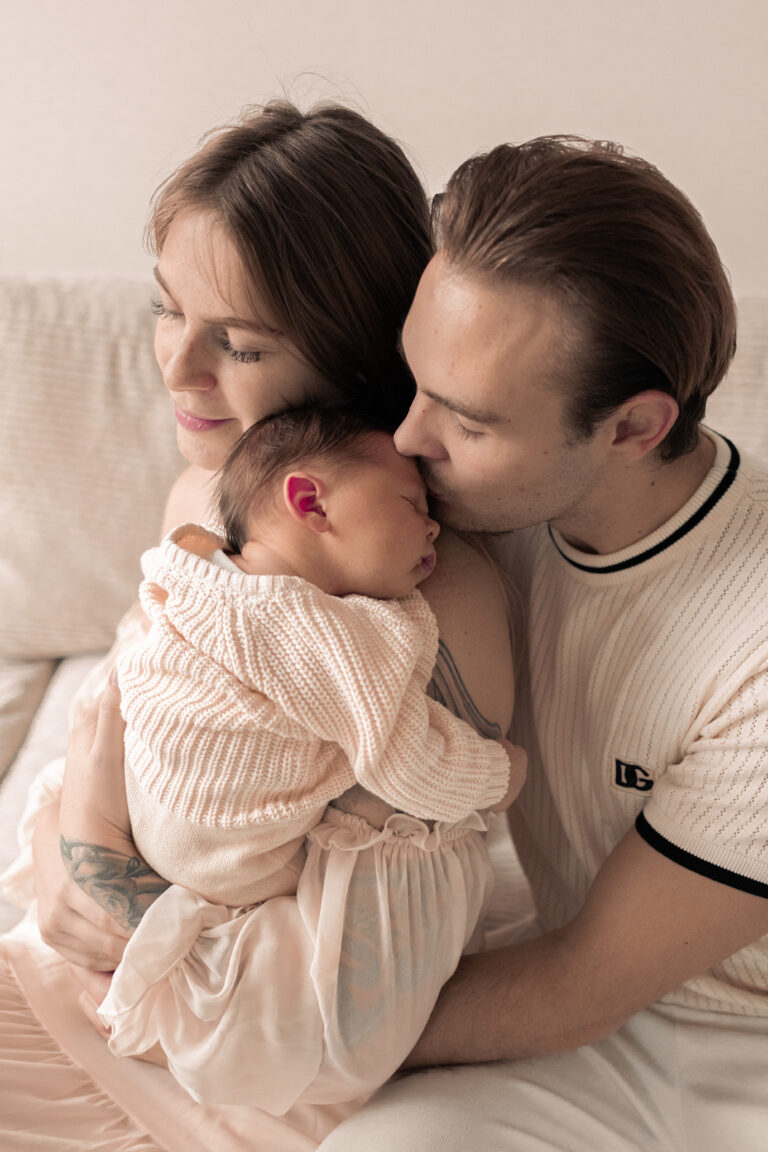 Frau hält Neugeborenes im Arm, sitzt auf Sofa, Mann umarmt sie und küsst das Baby.