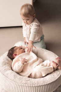 newbornshooting-nuernberg Neugeborenes liegt auf Decke, ältere Schwester greift nach ihr im Wohnzimmer.