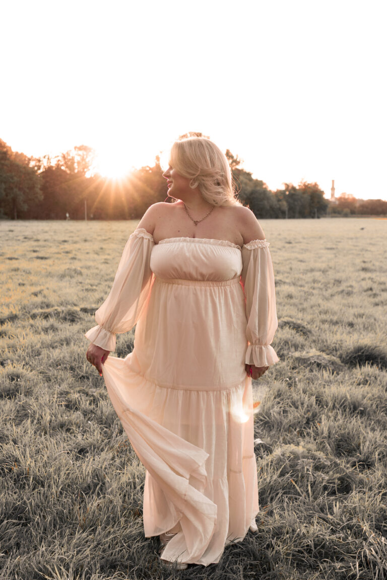 Blonde Frau auf Wiese im Sonnenuntergang trägt helles Kleid und blickt zur Sonne.