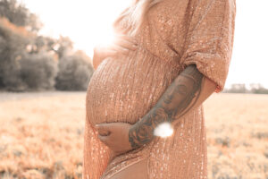 schwanger-shooting-outdoor-fuerth Schwangere Frau steht im goldenen Kleid vor dem Sonnenuntergang auf Wiese in Fürth und hält ihren Babybauch fest.