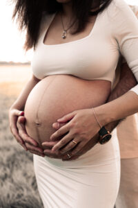 schwangerschaft-natur-fuerth Frau im weißen Kleid steht im Sonnenuntergang, ihr Mann steht hinter ihr und hält ihren Babybauch fest auf Wiese in Fürth.