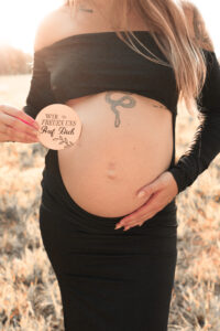 schwangerschaftsfotografie-fuerth Schwangere Frau hält ein Schild mit ‚Wir freuen uns auf dich‘ hoch und hält dabei ihren Babybauch fest auf Wiese in Fürth.